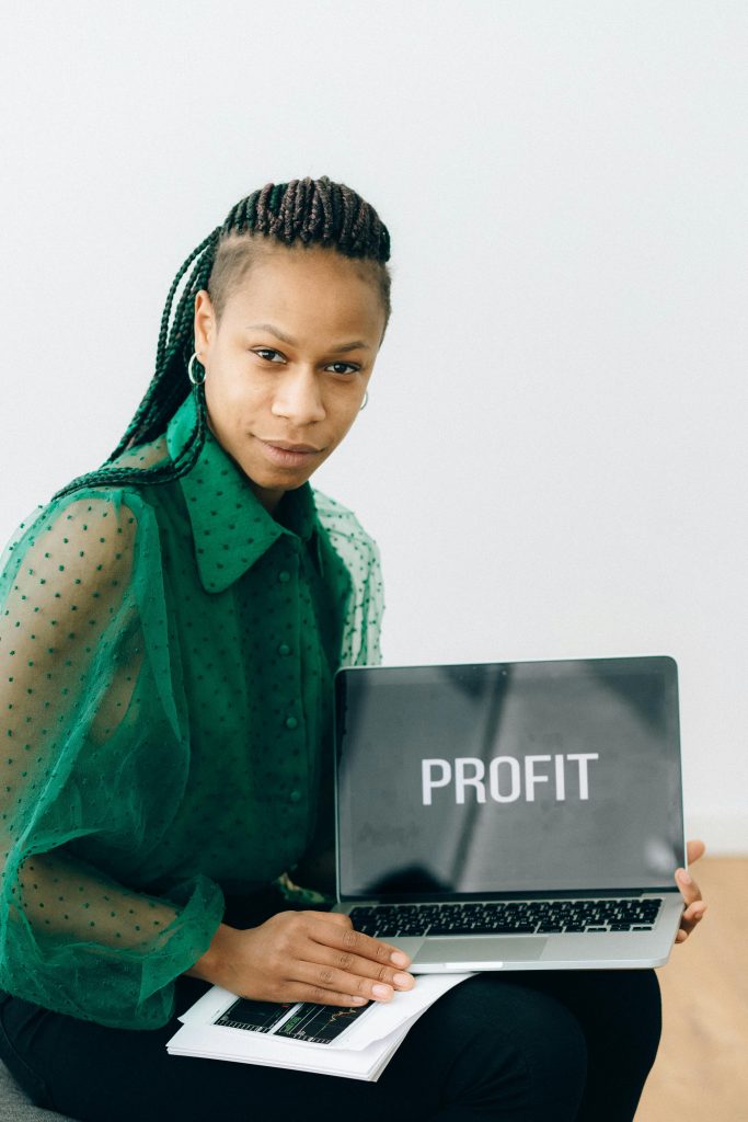 African American businesswoman in green blouse holding laptop with 'Profit' sign indoors.